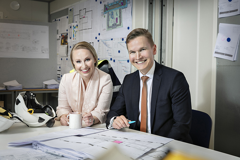 Anna ja Heikki pöydän äärellä, jossa rakennuspapereita ja kaksi valkoista kypärää.