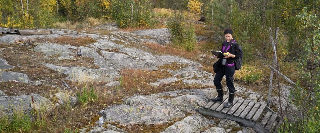 Nainen seisoo kalliolla kädessään karttoja ja paikanninlaite.