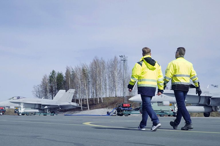 Kaksi miestä kävelee kiitoradalla, taustalla Boeing F/A-18 Hornet -monitoimihävittäjiä