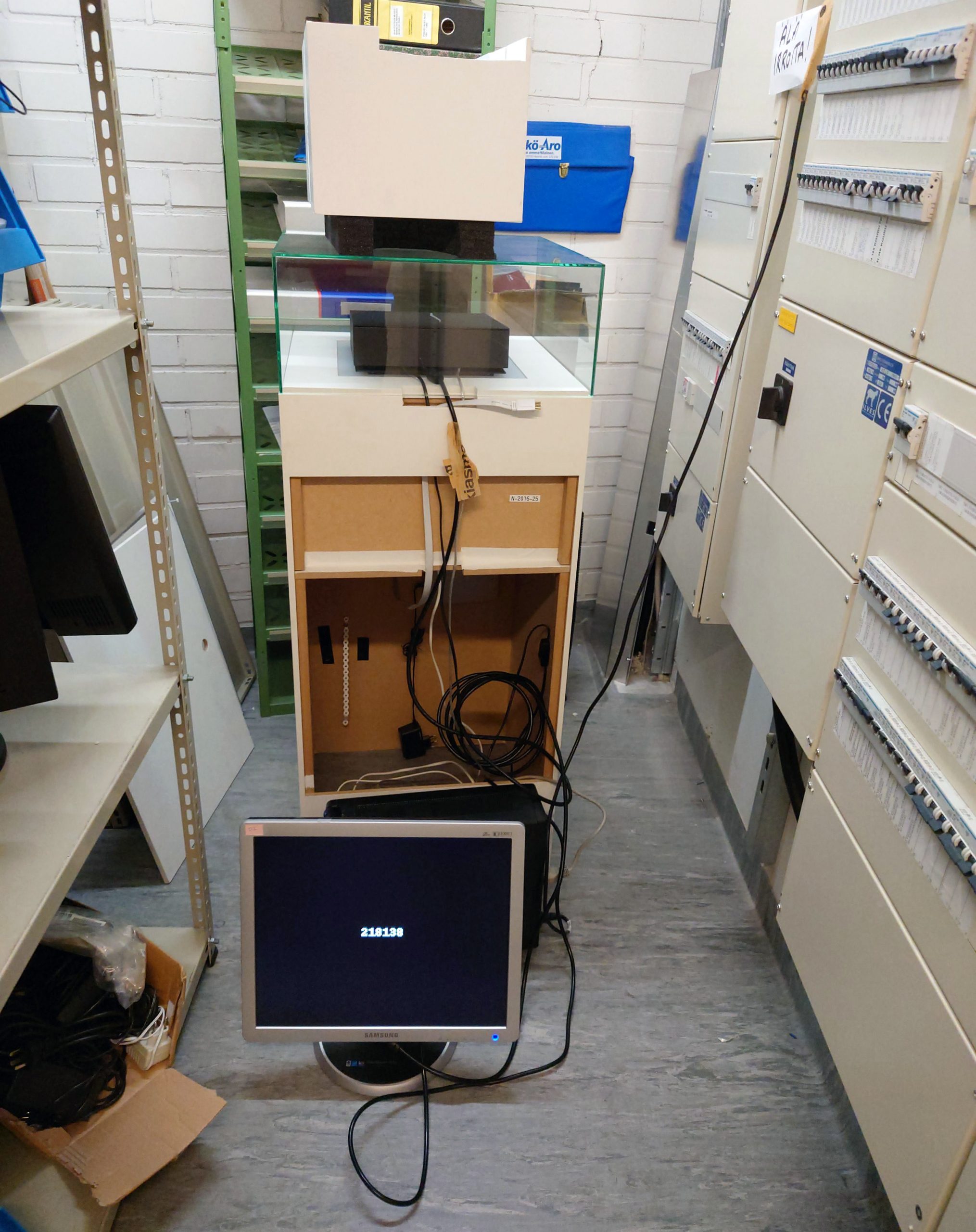 Harmaalla lattialla oleva tietokoneen näyttö, jossa musta tausta ja valkoinen numerosarja keskellä. Takana kaappi, jonka päällä lasivitriini, jonka sisällä musta laatikko. Lasivitriinin päällä on valkoinen laatikko. Oikealla sähkökaappeja ja vasemmalla varastohyllyjä.