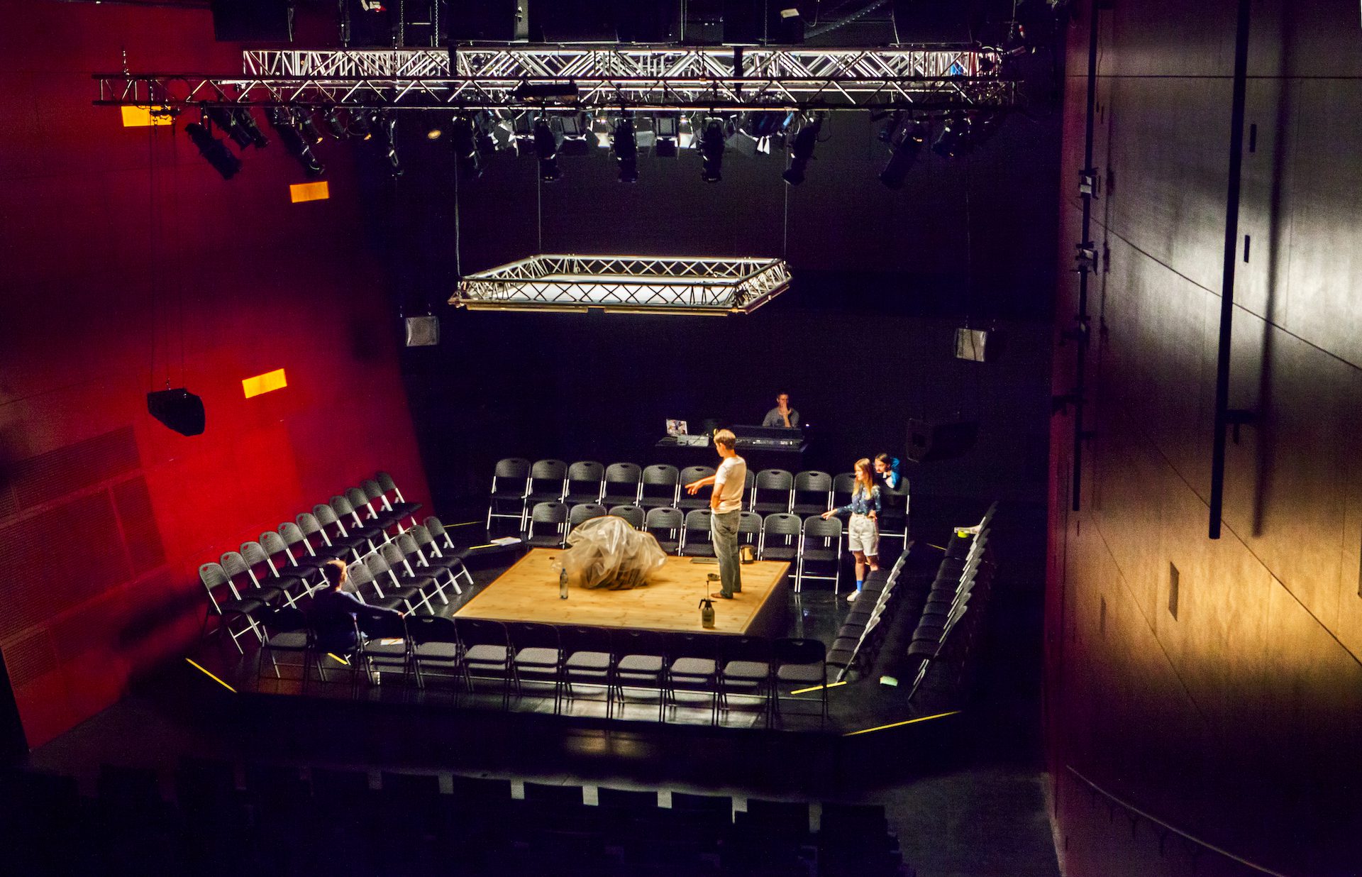 Neliö lava, jonka ympärillä on mustia tuoleja. Lavalla seisoo yksi henkilö ja lavan ympärillä neljä henkilöä. Katosta roikkuu telineitä, joissa on kiinni valoja. 