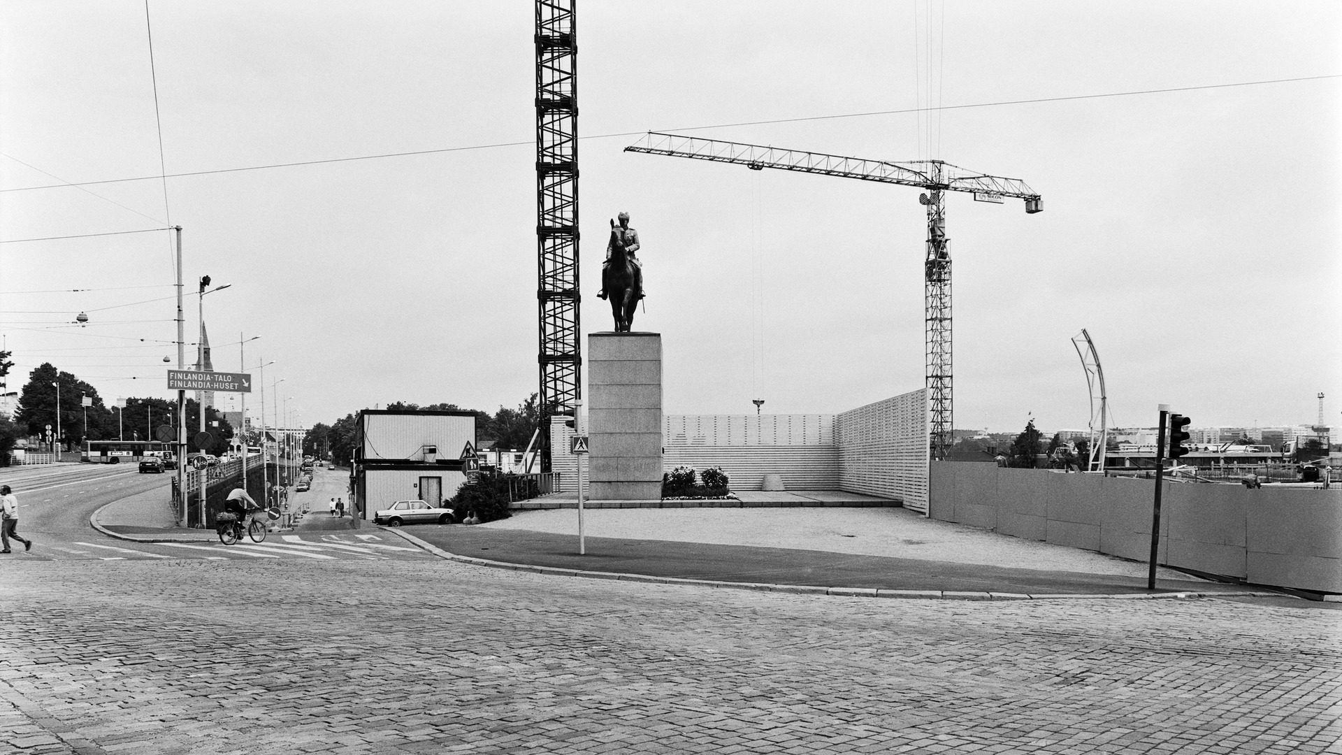 Mustavalkoinen kuva, jossa näkyy Mannerheimin ratsastajapatsas, matala työmaa-aita ja kaksi nostokurkea. Mannerheimintie on kuvassa liki autio, vain yksinäinen jalankulkija ja pyöräilijä näkyvissä, kaukana Eduskuntatalon kohdalla näkyy auto.