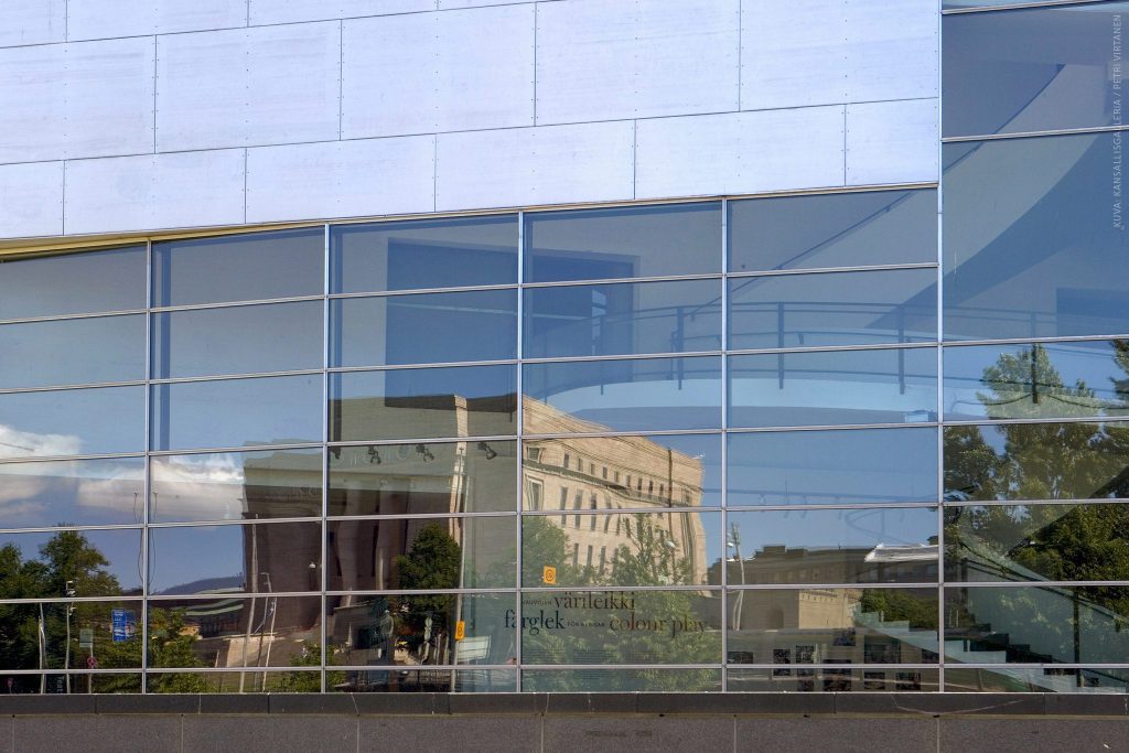 Valkoista seinää ja lasi-ikkuna, johon heijastuu eduskuntatalo ja sininen taivas.