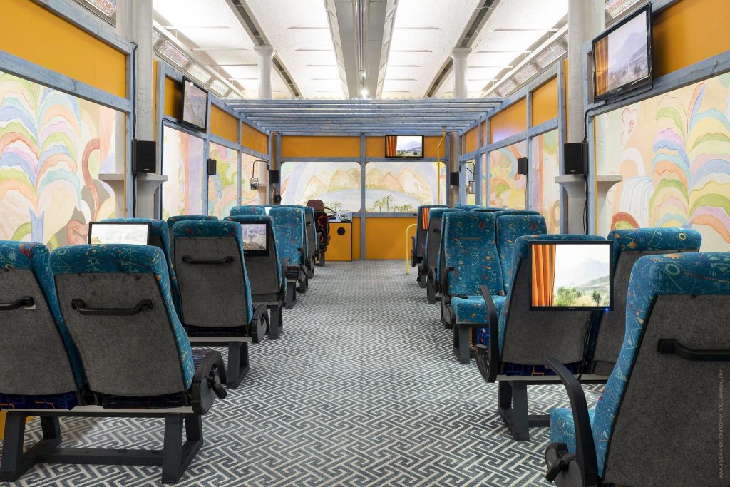 Bussin sisäosa, jossa on sini-vihreät kahden penkin rivit kummallakin puolella. Penkkien selkänojissa ja korkealla seinillä on televisioita.
