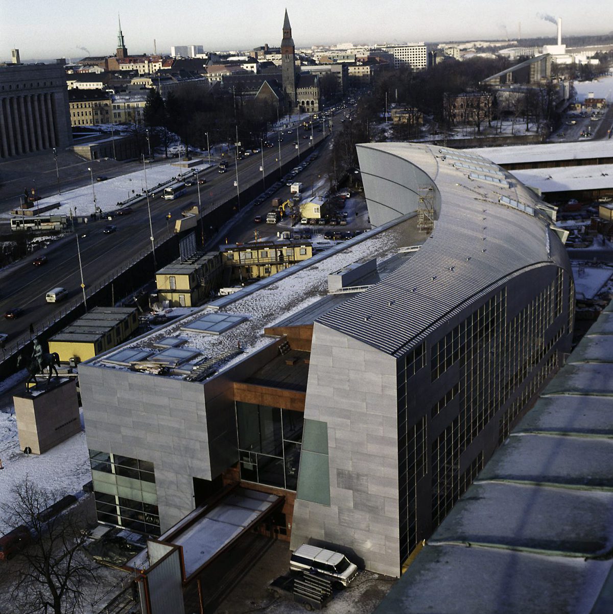 Kiasma kuvattuna etelästä. Harmaat seinät ja katto, seinissä myös punaruskeita kohtia. Ikkunoita seinissä ja katossa. Katolla lunta, tikkaat ja rakennusmateriaaleja. Taustalla eduskuntatalo ja Kansallismuseo.