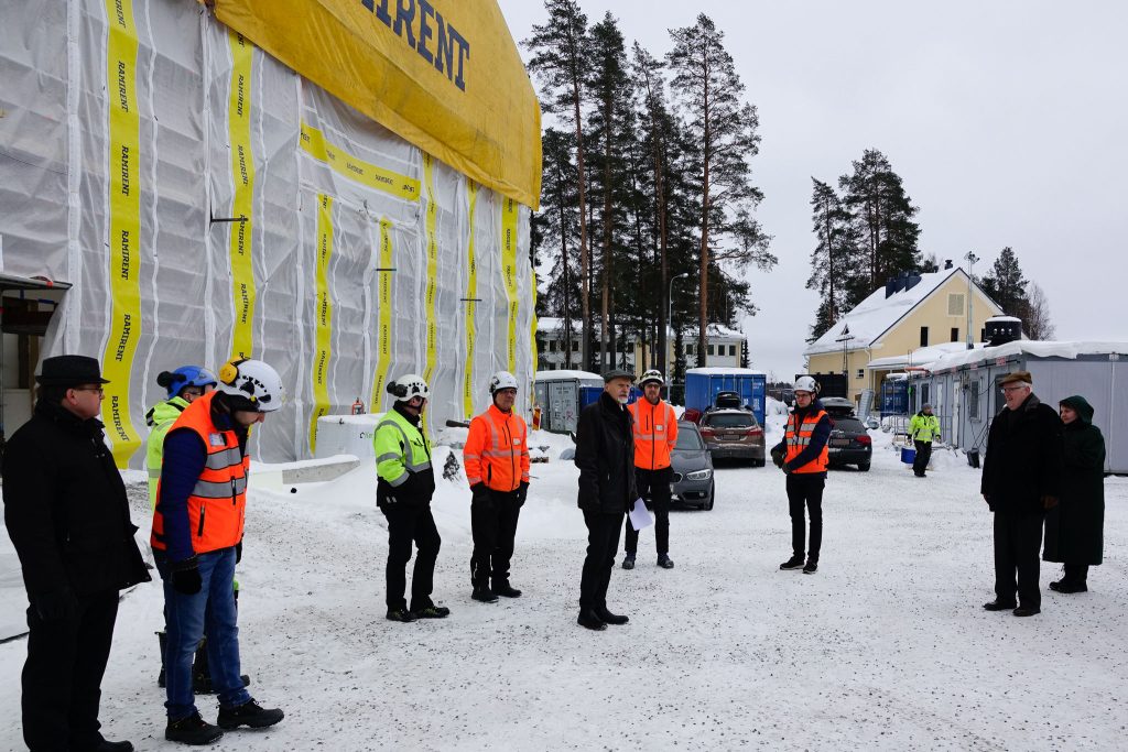 Rakennustyömaalla kelta-valkoisen sääsuojan alla sotilaskoti-liikuntarakennus, jonka edessä ihmisiä. Työmaakoppeja taustalla oikealla.