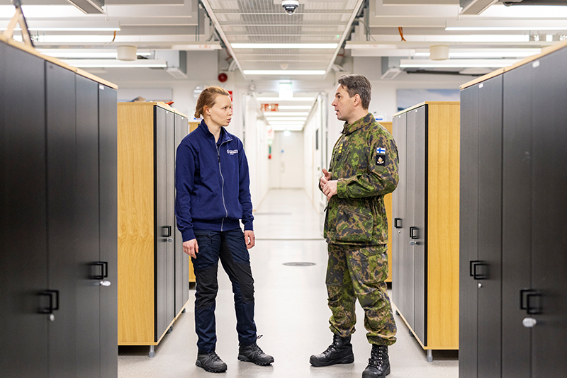 Sinitakkinen Puolustuskiinteistöjen asiantuntija ja Puolustusvoimien virkahenkilö keskustelevat.