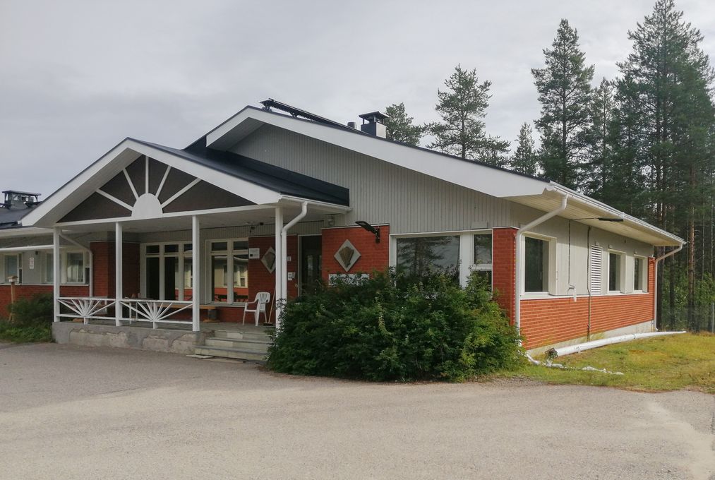 Punatiilinen talo, jossa valkoinen katto ja kuisti, ympärillä metsä.