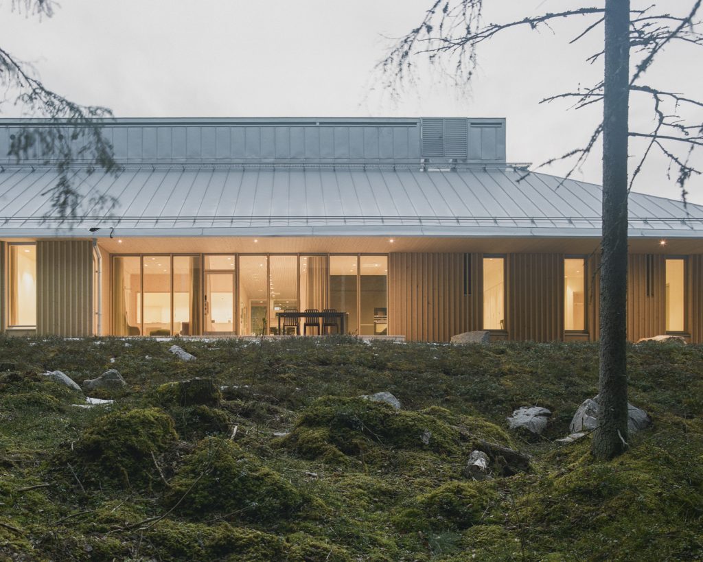 Metsämaisema, jonka takana puurakenteinen rakennus. Rakennuksessa kaunis valaistus.