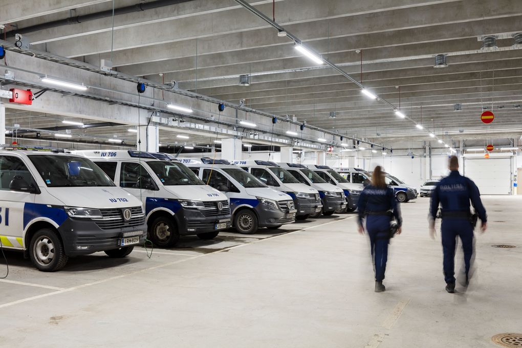 Rivi poliisiautoja pysäköitynä autotallissa, kaksi ihmistä kävelee poispäin.