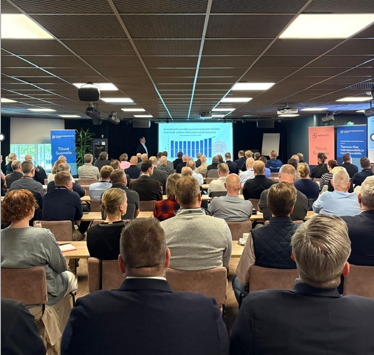 Salissa istuu runsaasti ihmisiä kuuntelemassa puheenvuoroa hankinnoista.