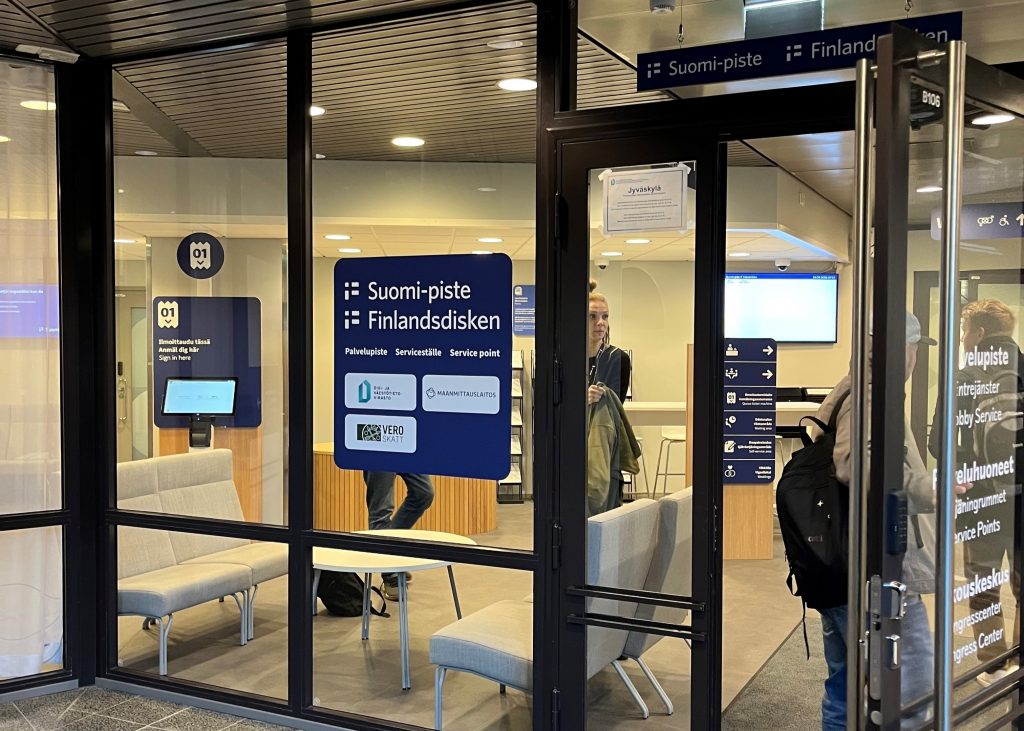 Sisäänkäynti Suomi-pisteeseen, lasiovien takana näkyy odotustila ja ihmisiä.