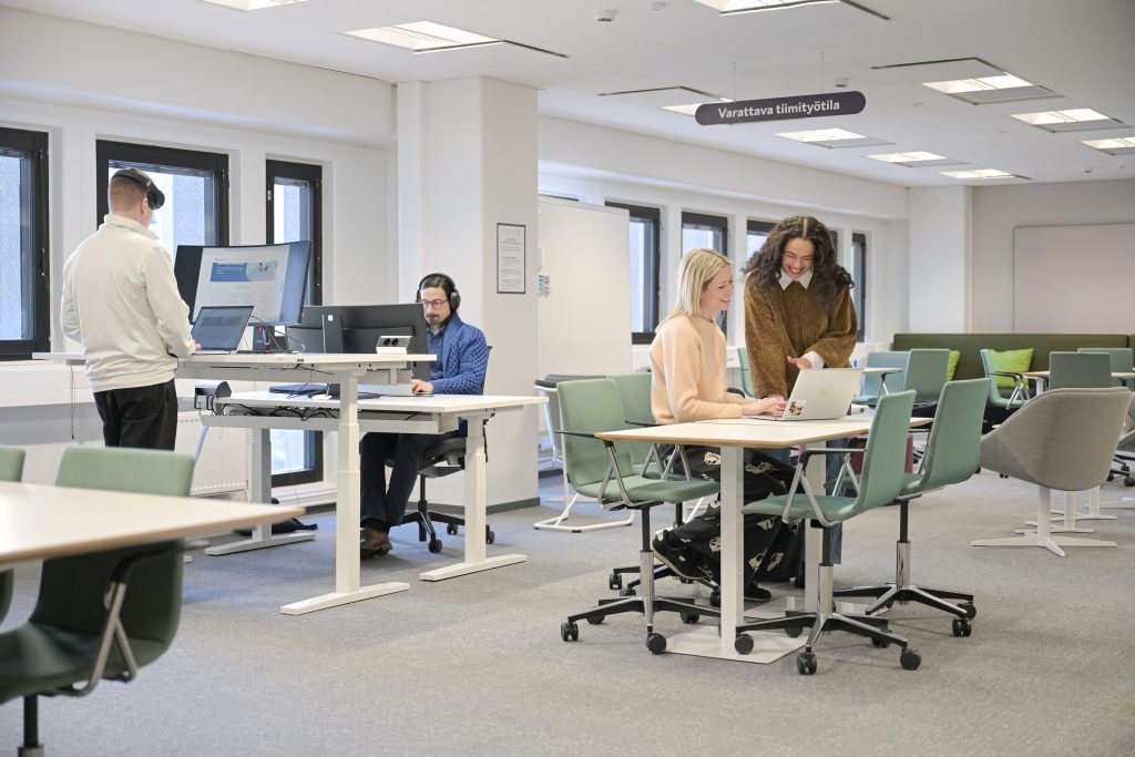 Työntekijöitä yhteisessä työympäristössä. Katossa kyltti "Varattava tiimityötila".