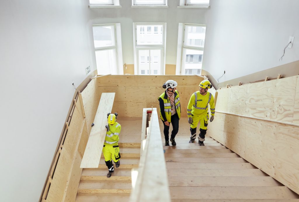 Kaksi henkilöä kävelee ylöspäin talon sisällä olevan työmaan portaita. Kolmas henkilö menee portaita alaspäin ja kantaa olallaan lautaa.