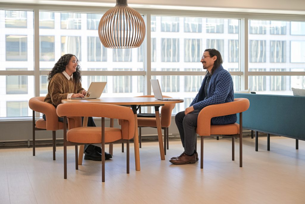 Kuvassa yhteisessä työympäristössä kaksi henkilöä pöydän ääressä keskustelemassa tietokoneet edessään.