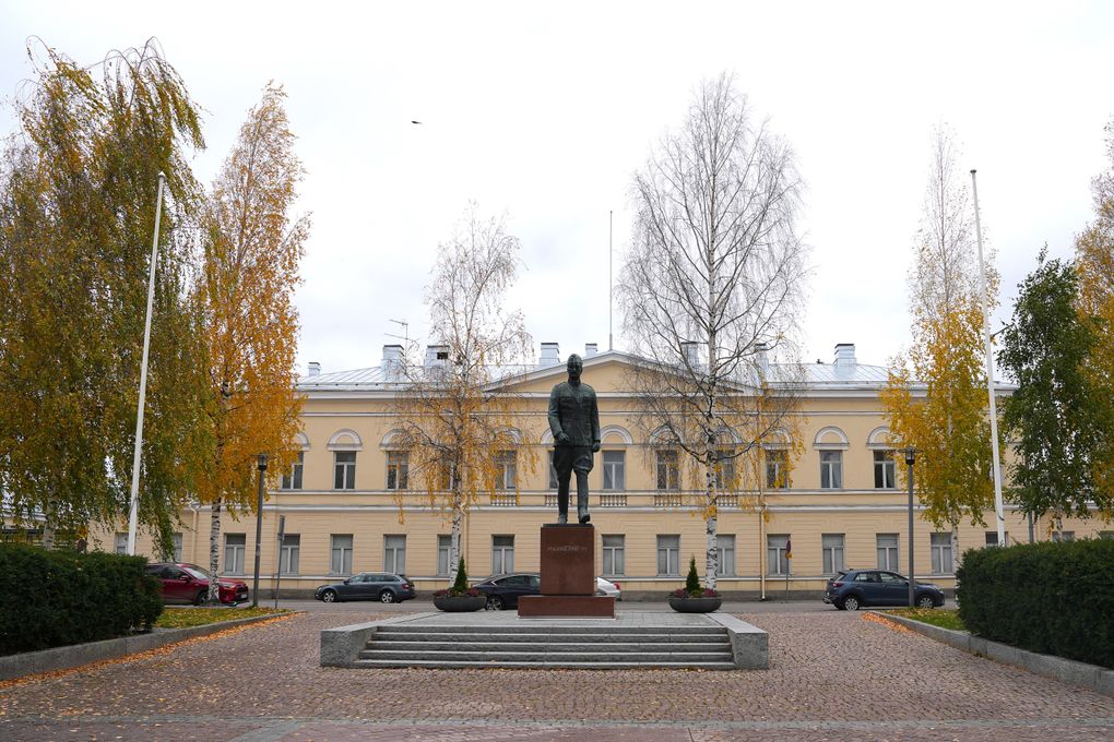 Patsas aukion keskellä syksyisessä ympäristössä, takana vaalea rakennus ja puita.