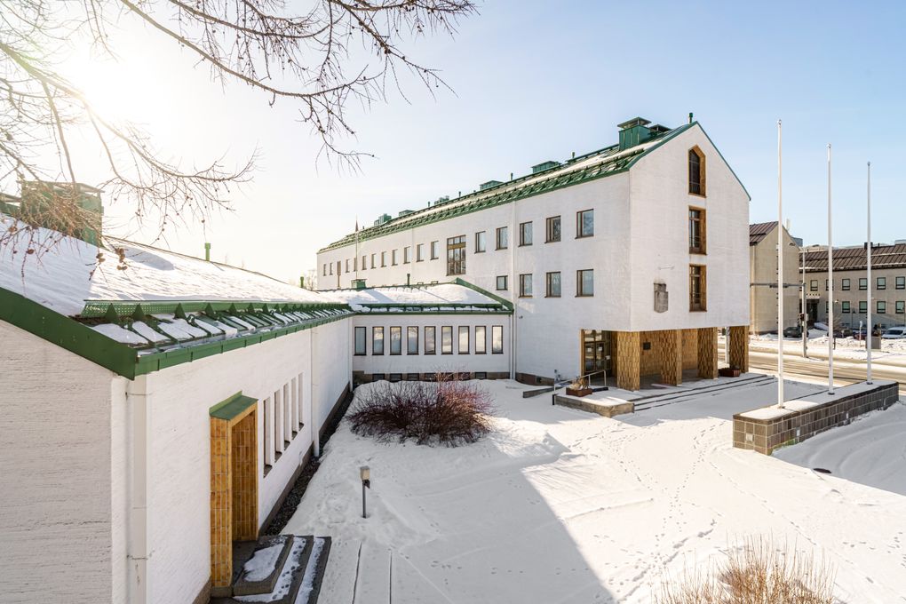 Valkoinen, moderni rakennus lumisessa maisemassa. Aurinko paistaa kirkkaalta taivaalta.