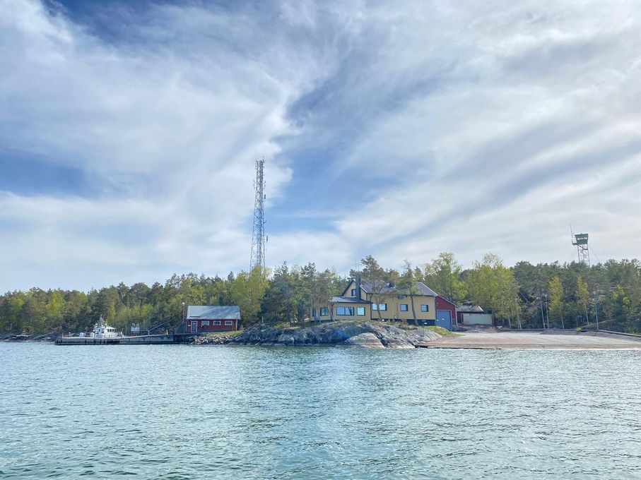 Mereltä kuvattu saariston rannikkokohde, jossa vaalea päärakennus, punaisia talousrakennuksia, laituri ja korkea masto kallioisella, metsäisellä saarella. Taivaalla pilviä.