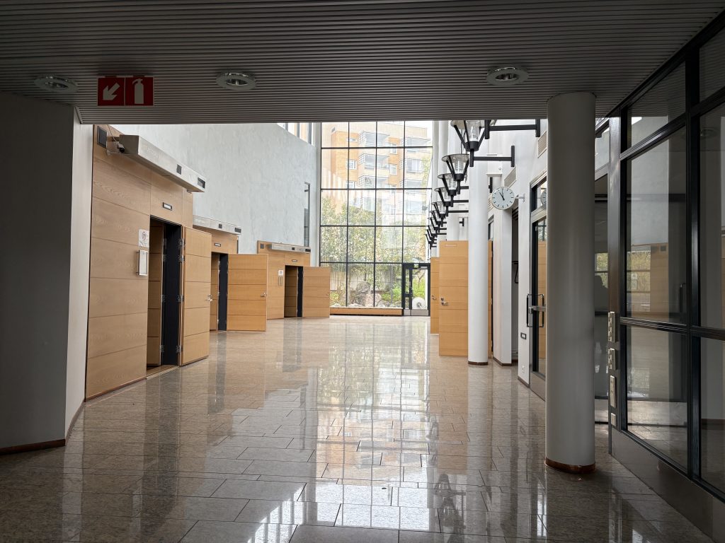 Rakennuksen läpileikkaava valoisa iso ala-aula. Valkoinen laatoitettu lattia ja päädyssä lattiasta kattoon ulottuva lasiseinä.