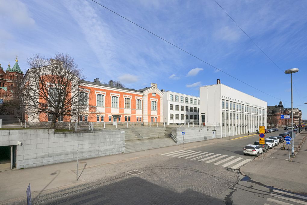 Kuva näyttää kadun, jonka vasemmalla puolella on oranssi ja valkoinen rakennus. Oikealla on moderni valkoinen rakennus. Kadulla on pysäköityjä autoja ja suojatie. Taustalla on sininen taivas.