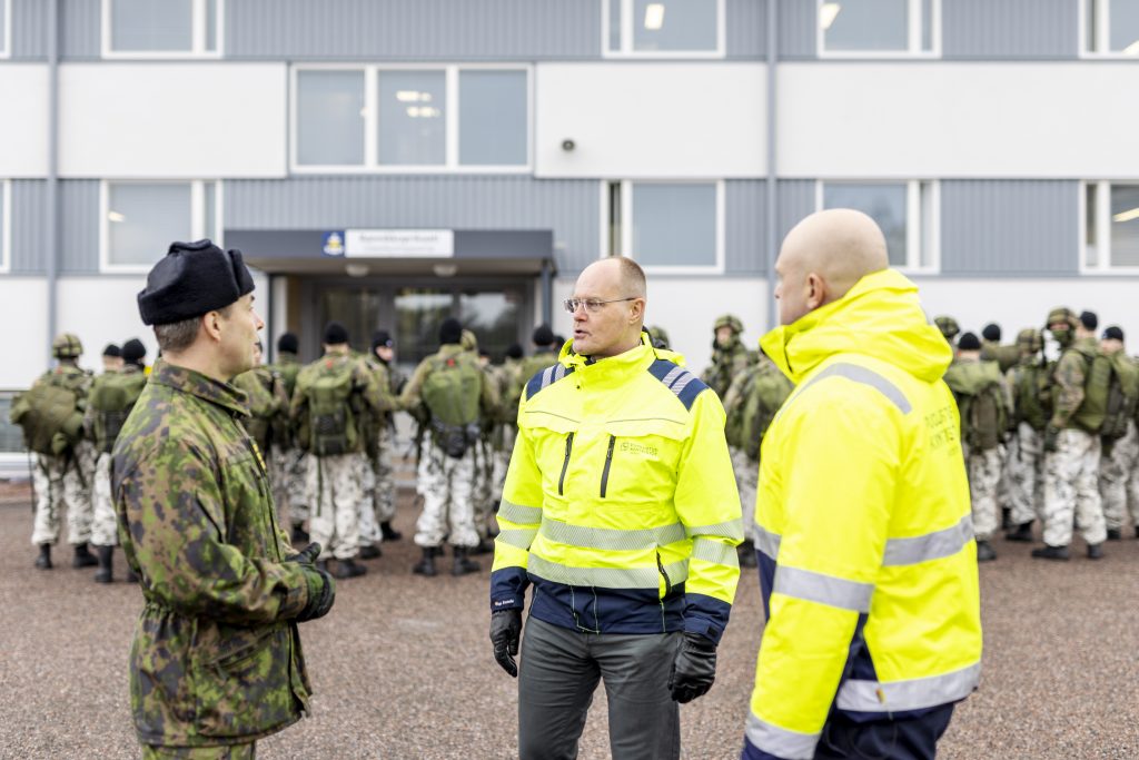 Kaksi Puolustuskiinteistöjen työntekijää keskustelee Puolustusvoimien työntekijän kanssa. Taustalla seisoo varusmiehiä kasarmirakennuksen edessä.