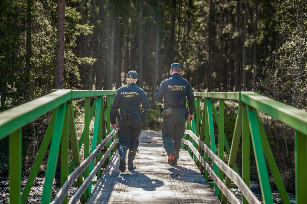 Kaksi henkilöä kävelee vihreällä sillalla metsässä. Heidän univormuissaan lukee "RAJAVARTIOLAITOS".
